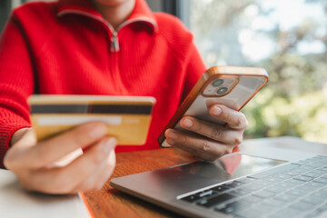 Close-up of a person using a smartphone and credit card for online shopping, sitting at a table with a laptop.