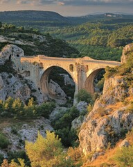 Obraz premium Ancient stone bridge spanning a deep valley at sunset, surrounded by lush greenery and rocky terrain.