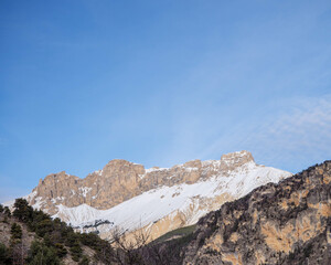 Snowy pic de bure and orange cliffs rise over evergreen trees blue sky