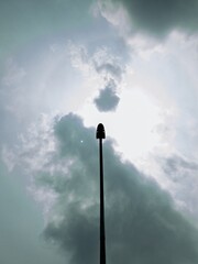 Silhouette of a curved streetlight against a cloudy sky with sunlight filtering through. A moody and dramatic scene showcasing urban minimalism and contrast between light and dark.
