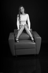 Fototapeta premium Long legged young woman in white dress sitting on a chair black and white low key portrait