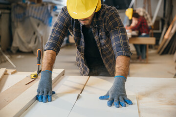 Young carpenter in a yellow helmet, blue gloves, and plaid shirt using a circular saw to cut wood in a furniture factory. Ideal for showcasing woodworking skills and construction techniques. Labor day