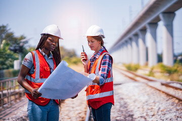 Black and White female Rail transportation engineers in safety vest and hardhat check the...