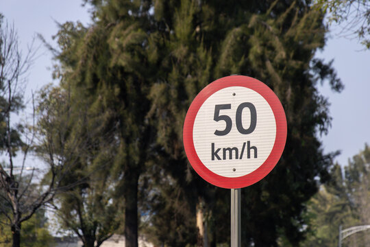 50 km per hour speed limit sign with a red border stands on a roadside, surrounded by trees and natural scenery in daylight.