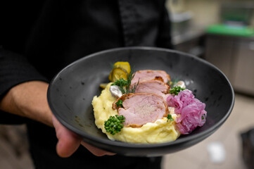 A chef presents a gourmet plate featuring smooth mashed potatoes, tender pork slices, pickles, and colorful garnishes, showcasing culinary artistry in an elegant setting