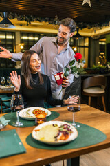 Boyfriend behind his girlfriend holds rose and gift and he is surprising her on a romantic dinner date in restaurant.