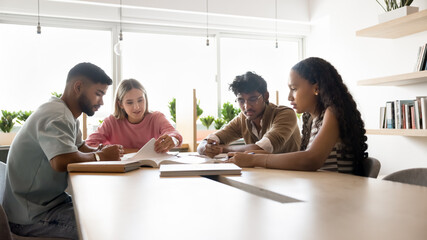 Four students gathered around table in modern study space, engaged in collaborative learning session, discussing their work, taking notes, exchanging ideas, debating topics, working on joint project