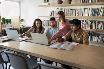 Four focused multiethnic students do assignment in library, discuss online task on laptop, preparing for exam together, search information on internet, share ideas, working on project. Education, tech