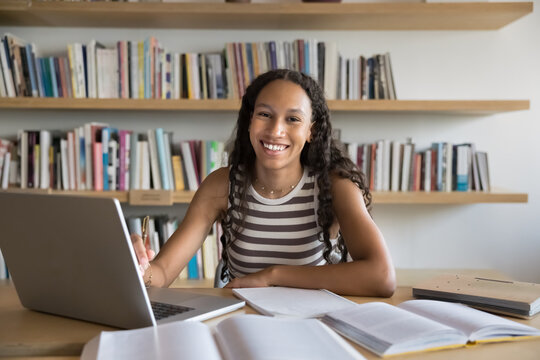 Portrait of schoolgirl do homework in library. Excellent student posing for camera with contented smile seated at desk with laptop and textbooks, enjoy effective e-learning using educational platform