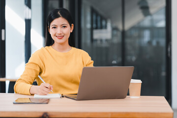 Fototapeta premium Woman in yellow sweater working on laptop while taking notes, sitting at wooden desk with coffee cup in modern office.