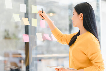 Woman in yellow sweater writes on colorful sticky notes on glass wall, brainstorming ideas.