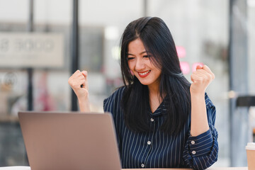 Cheerful Asian woman celebrating success while working on laptop in modern office.