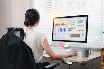 Asian businesswoman analyzing data on computer screen with charts and graphs in modern office.