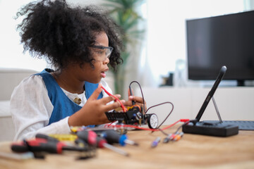 Black schoolgirl build electrical robot with concentrate by watching tablet. Smart girl fix her robotic car at home. Female student learn about electric robot desing.