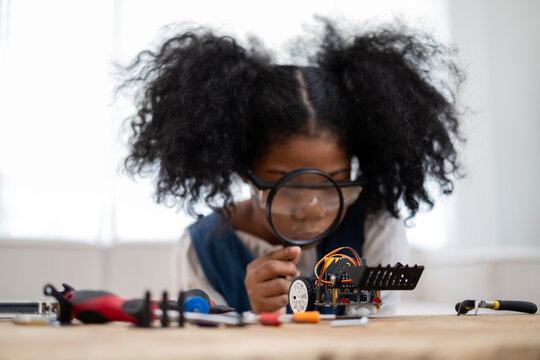 Black schoolgirl look through manifying glass to observe tiny pieces of robotic car. Smart girl learn to build robot.