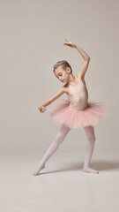Graceful kid, charming little ballerina dancer, dressed pink tutu and tiara, training dance moves against warm grey background. Concept of ballet, elegance and beauty, grace. Ad
