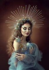 Young beautiful woman with long curly hair in transparent blue fabric and in a chic crown. Portrait of a Greek goddess. Mystical image. Decorative makeup. Glow effect. Fairytale image of a queen.