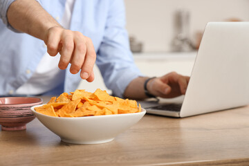 Man with laptop eating tasty nachos in kitchen
