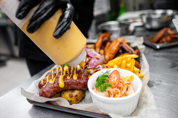 A chef in black gloves is drizzling sauce over a platter of grilled meats, potatoes, and fresh vegetables, showcasing appetizing barbecue cuisine in a vibrant kitchen setting.