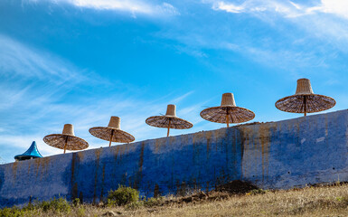  Wicker parasols lined up behind a blue wall, under a clear sky, capturing the Mediterranean essence of Chefchaouen, Morocco.