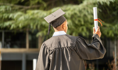 rear view of graduate afro american student boy raising diploma wearing cap and gown outdoors