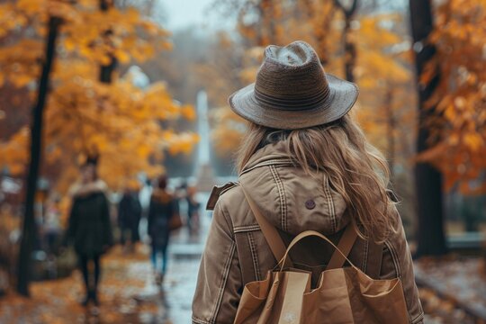 A woman with long hair and a hat strolls in a park filled with vibrant autumn leaves. People can be seen enjoying the scenery nearby, with a fountain in the background