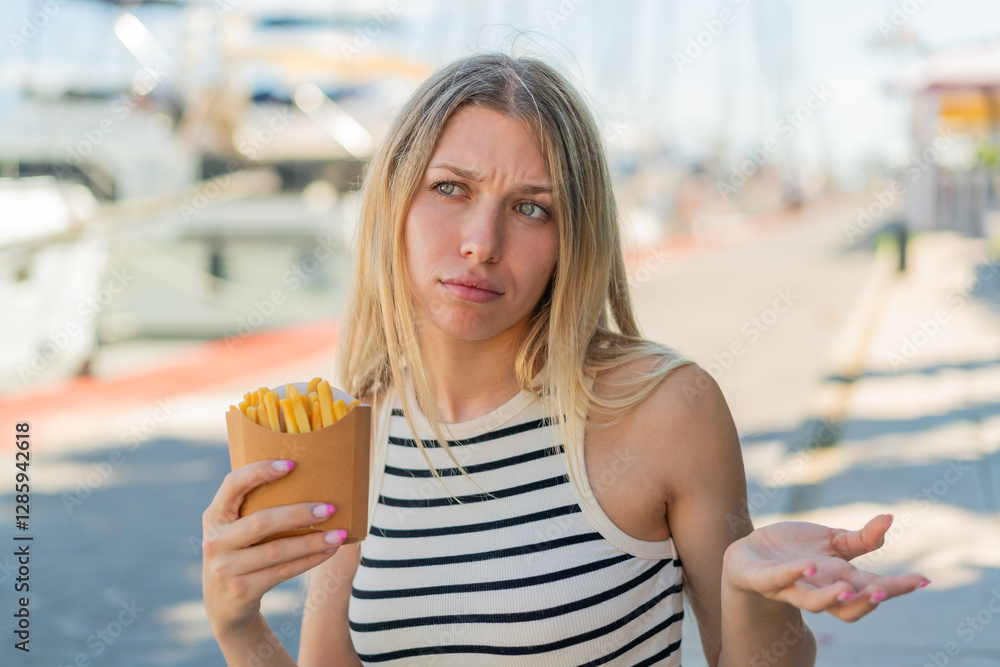 Wall mural Young blonde woman holding fried chips at outdoors making doubts gesture while lifting the shoulders