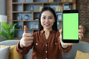 Asian woman smiling, holding smartphone with green screen, showing thumbs up, sitting on couch in home setting. Concept of technology, positivity, online communication.
