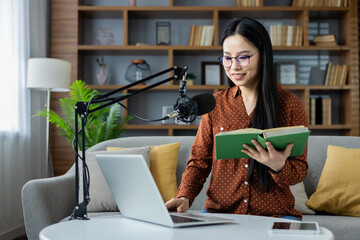Asian woman hosting podcast from home, using microphone, laptop, and book. She sits in comfortable,...
