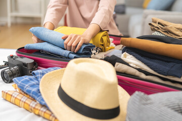 Travel concept, happy asian young woman hand preparing packing luggage bag for summer vacation trip on bed at home, pack her clothes and stuff, objects into bag suitcase for a new journey for leisure.