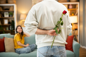 A man hides a red rose behind his back, ready to surprise a woman sitting on a couch, capturing a joyful moment.