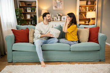 Obraz premium A woman is playfully surprised at her birthday gift while her partner shares the joy on a comfy couch in a cozy living room.
