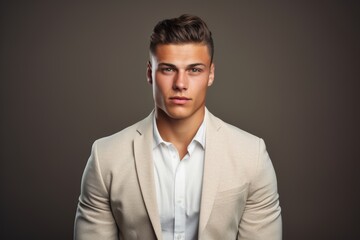 A man in a white shirt and tan jacket is standing in front of a wall