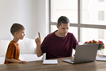 Focused self-employed father working on laptop while his little son asking attention, disturbing,...