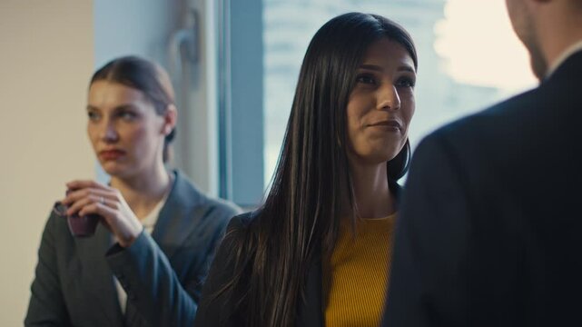 Professional woman in a suit holding a coffee cup observes a conversation between colleagues with a displeased expression. Concept of workplace dynamics, jealousy, and office rivalry