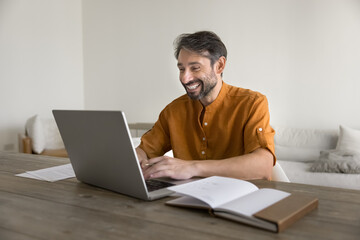 Happy middle-aged man sitting at table using laptop, smiling, reading pleasant news in notice or email, leading online chat, exchanging messages, working, spend time on internet during day-off at home