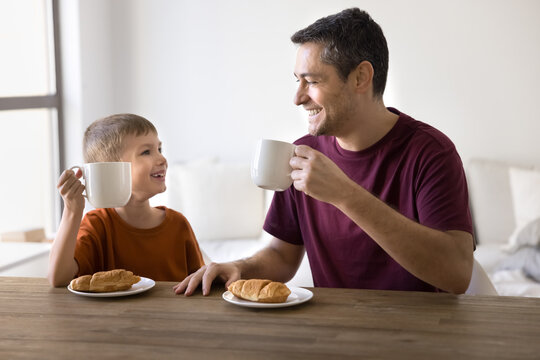 Father and little son sitting at table, holding mugs, enjoying breakfast with croissants and hot drinks, laughing, relish talk and time together. Family mealtime, morning rituals, parent-child bonding