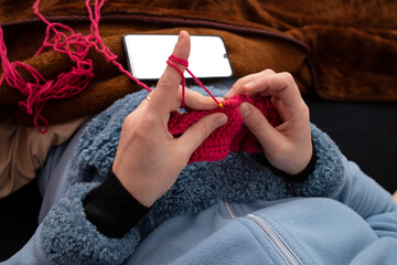 Woman hands knitting with a crochet hook and red thread , learning with a smartphone tutorial, soft...