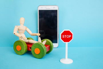 wooden mannequin on a toy car next to a defocused smartphone with a stop road sign on blue background, soft focus close up