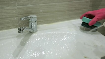 A woman in gloves sprays a special detergent on a chrome faucet and sink in order to clean it.