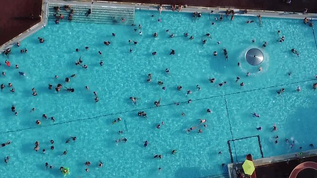 Top down drone footage of a group of people swimming in a public pool in the park on a sunny day