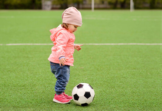 Little toddler girl in pink jacket, hat and sneakers playing with soccer ball at football field or stadium outdoors. Green background. Child dreams, girl power and active childhood concept. Copy space