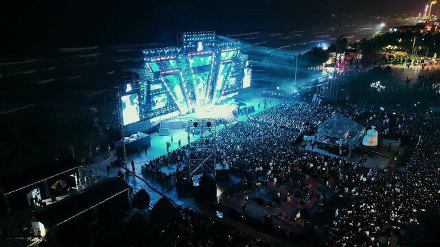 Open air music concert festival. Massive crowd gathering for night event near coastline. Colorful stage lighting illuminating energetic music performance with dynamic visual effects