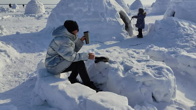 Adult happy woman relaxing on picnic in Eskimo village. Woman in warm down jacket near Igloo.