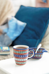 Ceramic mugs with brown ornament with tea on the kitchen table. There is a blue pillow on the sofa in the background. Selective focus. 