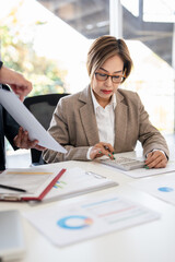 Senior professionals work on project in modern office, analyzing documents and charts. The man focuses intently the woman reviews papers with calculator ,dynamic scene of collaboration and expertise.