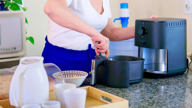 Woman using an air fryer on the countertop of a Brazilian home kitchen. She is placing breaded chicken drumettes into the appliance&rsquo;s basket to fry them