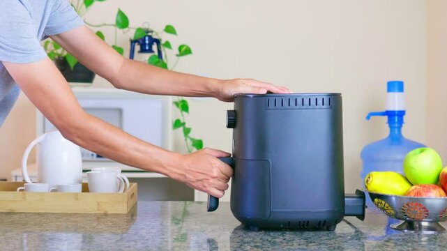 Hand placing the food basket into an air fryer. After inserting the basket with food, the person turns the dials to start the appliance. The air fryer is on a kitchen countertop