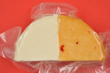 A top-down shot of a wedge of two-toned cheese, half white and half orange with red flecks, sealed in a clear plastic package against a red background