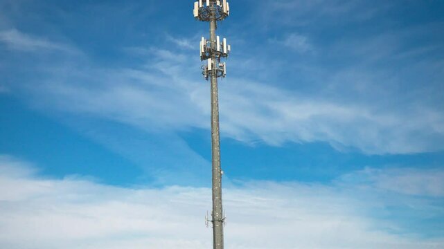 Tilting downward shot of a cellular communications tower and antenna providing digital service in a shopping area on a sunny day with clouds.